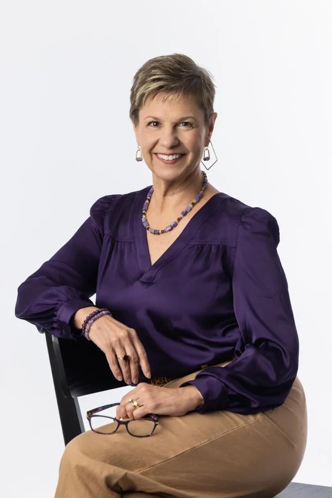 Professional studio headshot of woman seated and smiling wearing a purple shirt and holding glasses